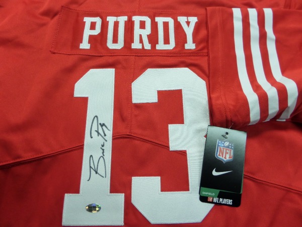 This red size L San Francisco 49'ers jersey from Nike is still tagged as new, and comes trimmed in white, with hand-sewn letters and numbers.  It is back number-signed in black sharpie by the emerging star QB of the Niners, Brock Purdy, and will look just great when framed for display.  Valued into the mid hundreds!