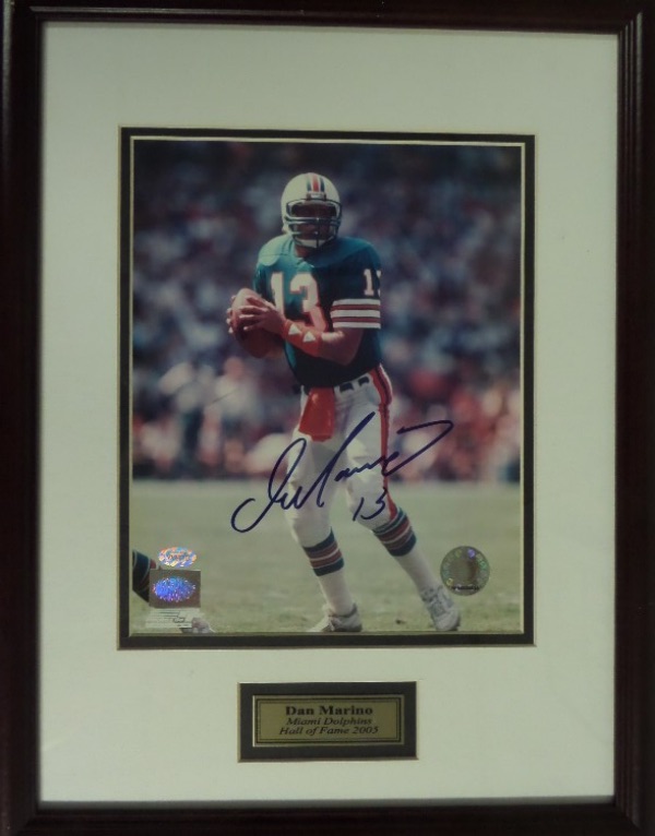 This 14x18 wood framed and double matted display features a custom nameplate and a color action photo of Dan Marino with the Miami Dolphins.  It is hand-signed beautifully in blue sharpie by the HOF passer himself, grading a 9 at least, and includes both a Mounted Memories COA/hologram and Marino's own hologram for rock solid authenticity.  Ready for display, and valued into the low thousands!